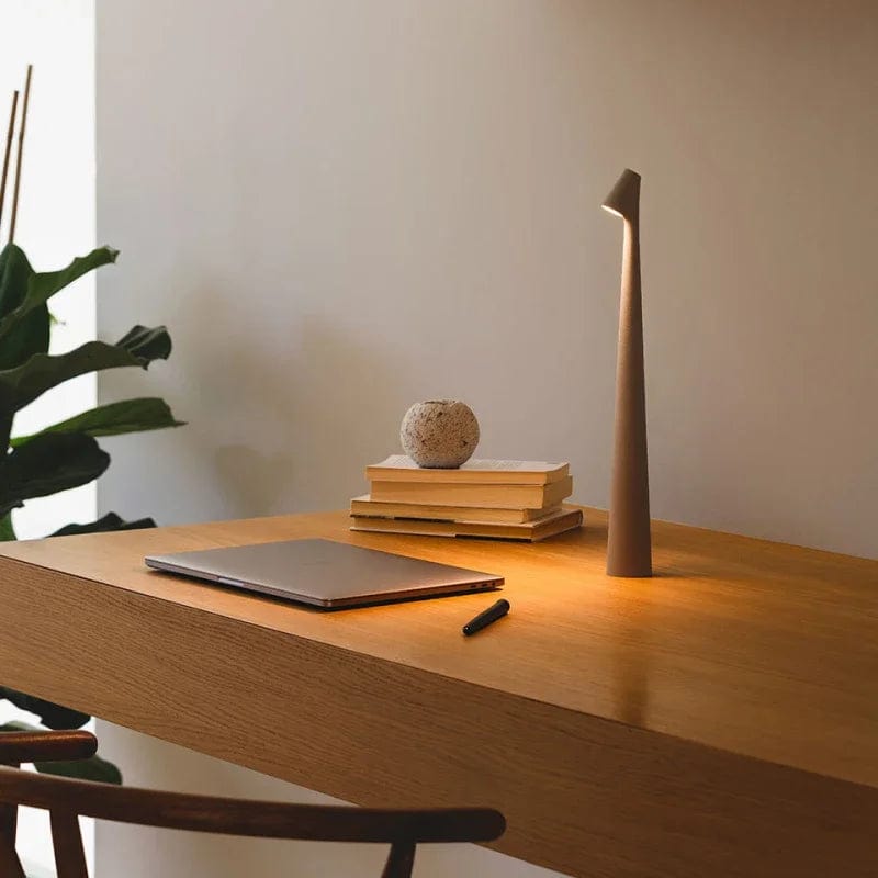 Grey Bexley table lamp lit on a wooden desk beside books and a laptop