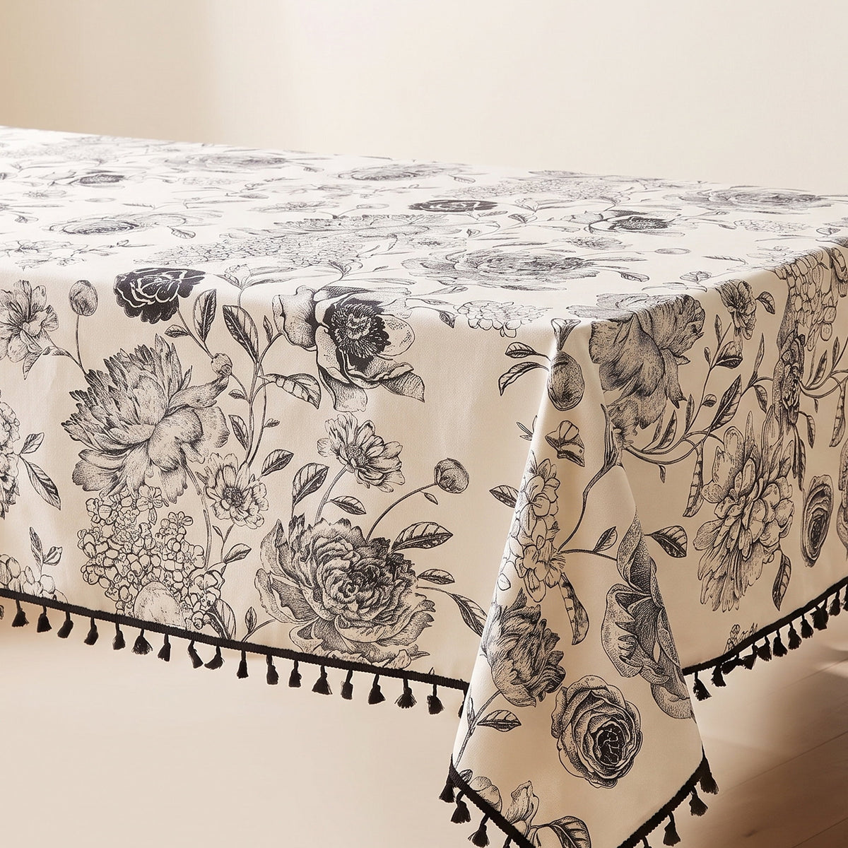 Blythe cream tablecloth with a black floral print and black tassel trim over a bare table, corner view