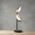 Cora bird table lamp unlit against a grey wall, two gold birds on a slim black post