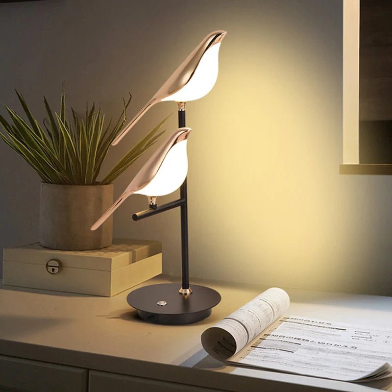 Cora bird table lamp lit on a sideboard beside a plant and a rolled drawing
