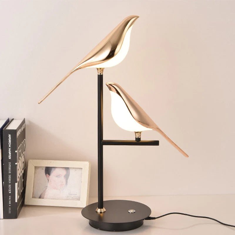 Cora bird table lamp lit on a wooden surface against a grey wall