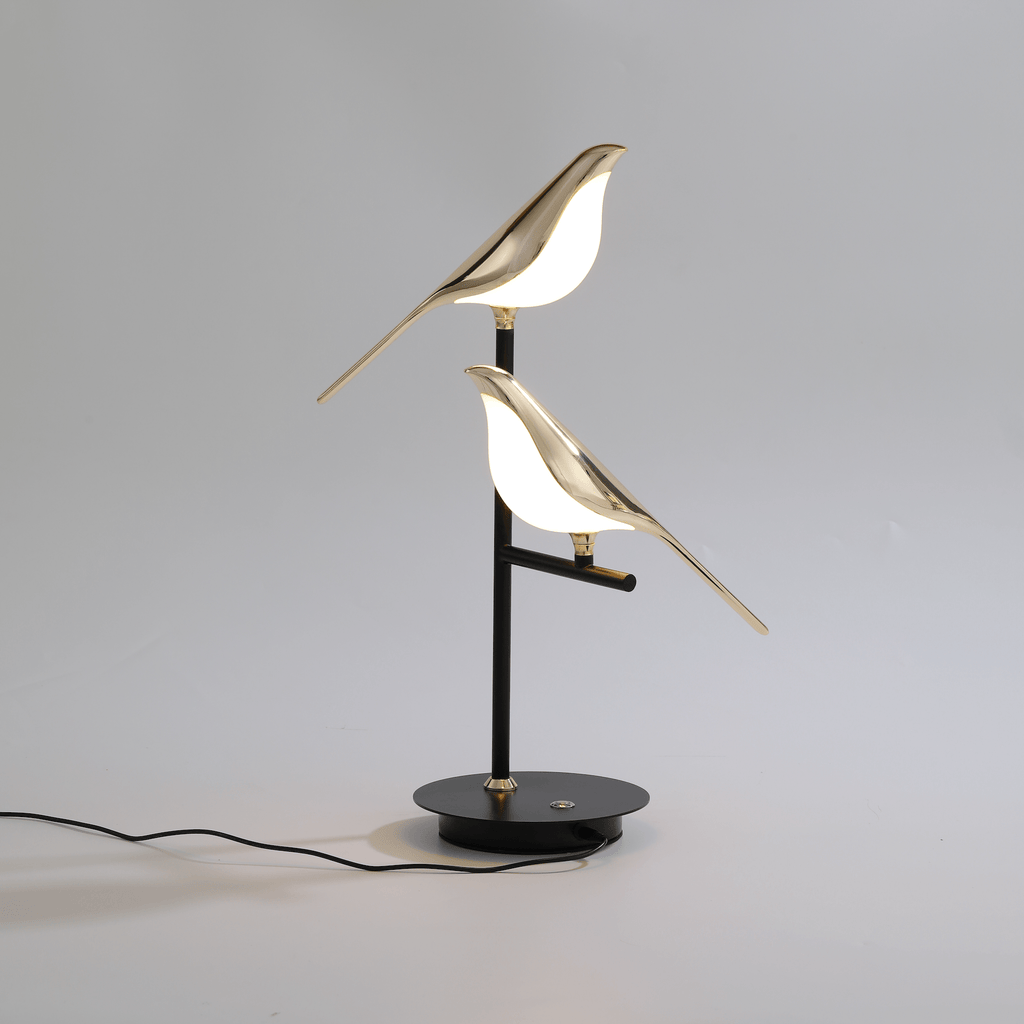Cora bird table lamp lit on a pale surface with the mains cable running from the base