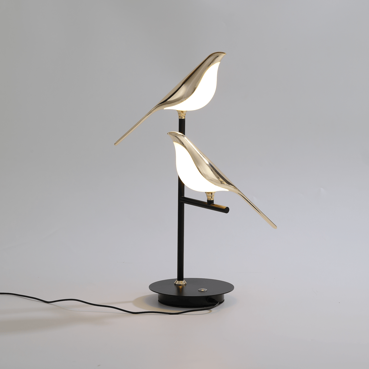 Cora bird table lamp lit on a pale surface with the mains cable running from the base
