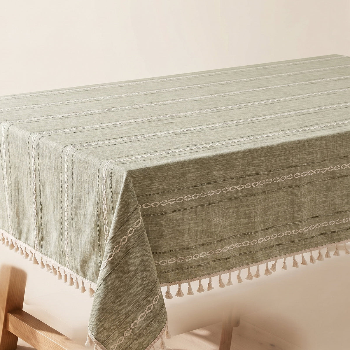Cora sage green tablecloth with striped embroidery and cream tassel edging over a bare table, corner view