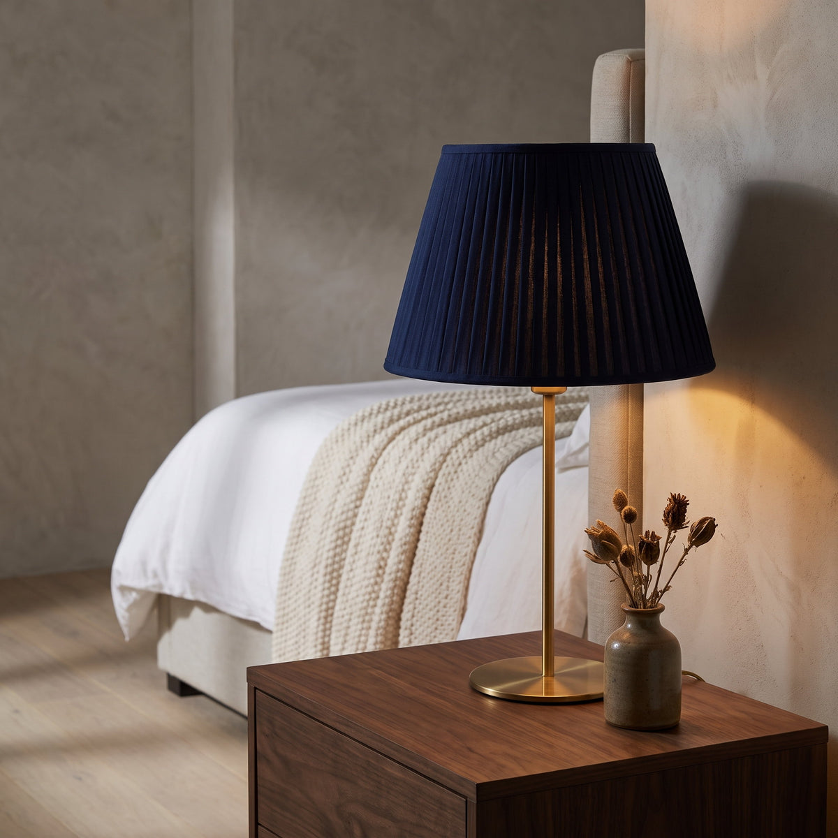Navy pleated table lamp lit on a walnut nightstand beside a linen bed, Style Room