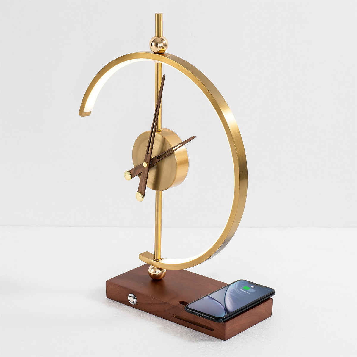 Orin clock lamp unlit, brass arc and analogue clock on a walnut base with a phone charging flat on the pad